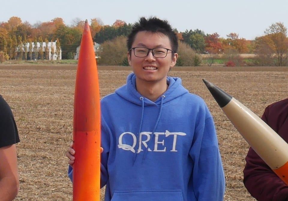 default-4738 A young man smiling while wearing a hoodie branded with QRET. He's holding a skinny orange rocket