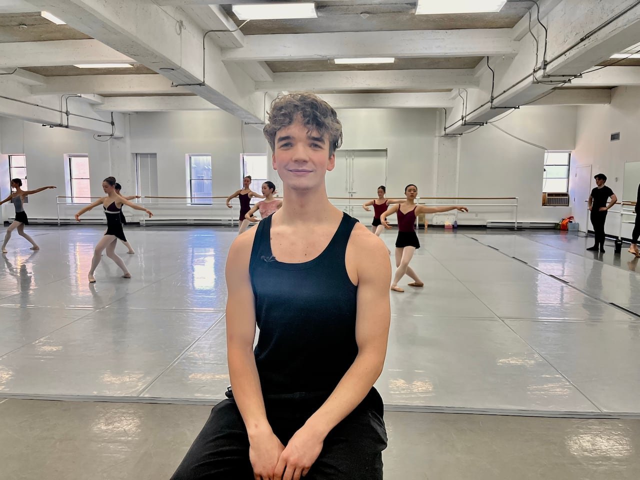 A ballet dancer sitting in a studio as others dance behind him.