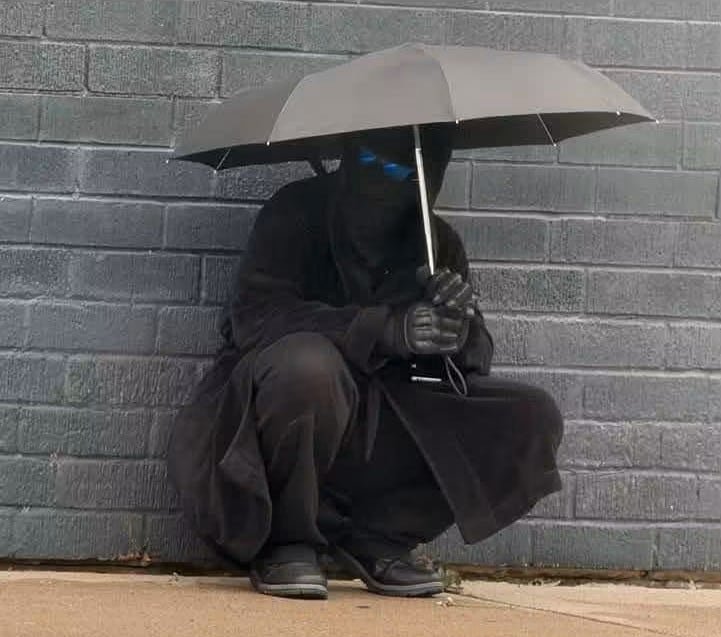 default-143 A man disguised in black attire, shown crouching on a sidewalk.