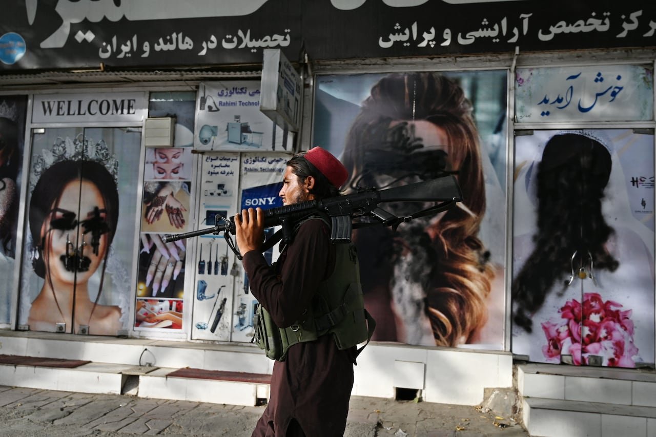 A man with a gun walks past a wall with women defaced using spray paint